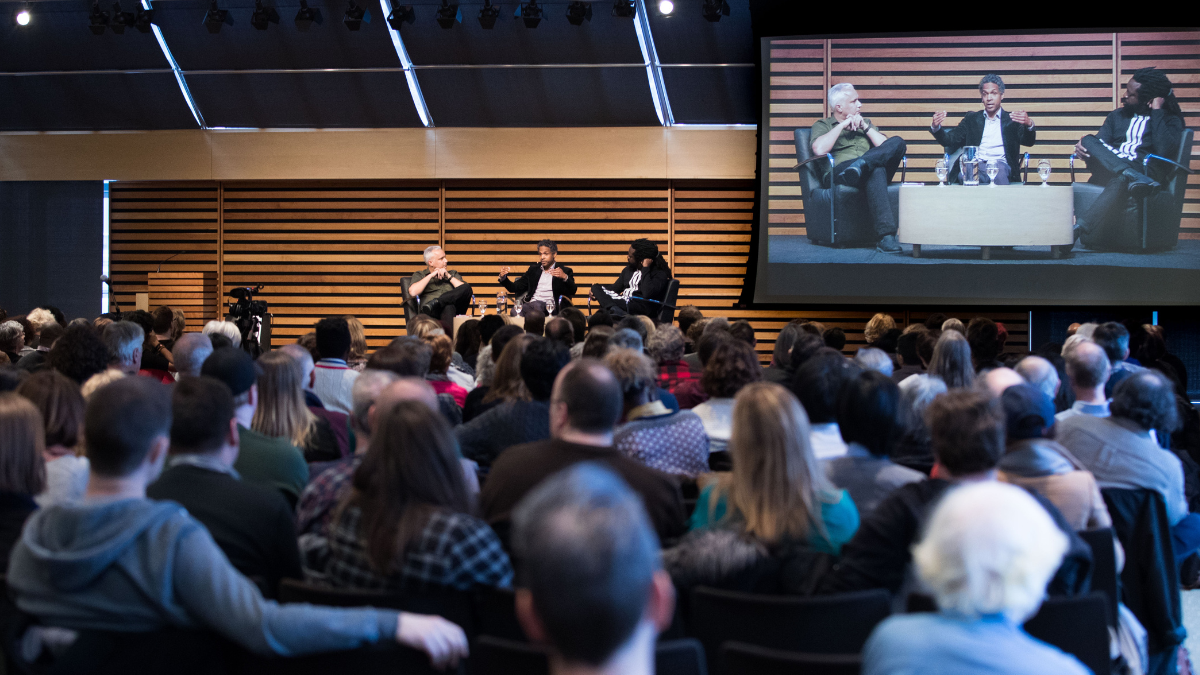 A seated audience watches a conversation on stage in the Appel Salon.