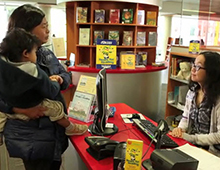 Parent hold child in arms in front the service desk at the library with a staff member behind the desk