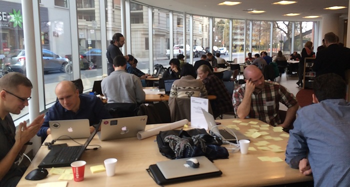 Open data hackathon, where a number of tables are placed around a well-lit round room with large windows running along the length of the wall. Outside the windows is a modern city streetscape. The tables in the room are large and seated at each table are 4 to 5 people working on their computers. The table in the foreground has people thinking and discussing, with their computers placed in front of them with sticky notes covering one side of the table. Coffee cups and a rolled up black jacket are also on the foreground table.